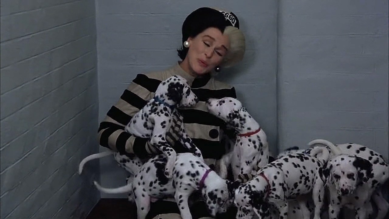 Glenn Close as Cruella in 102 Dalmatians sits in a bare jail cell wearing a black-and-white striped blouse and hat and cooing over six Dalmatian puppies in the live-action movie 102 Dalmatians