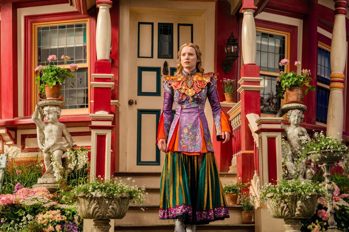Alice (Mia Wasikowska) stands in an absurdly brightly colored striped skirt and silk blouse with a humongous gold and red collar piece, in front of a similarly bright red-and-white painted-lady style house surrounded by greenery in Disney’s live-action movie Alice Through the Looking Glass