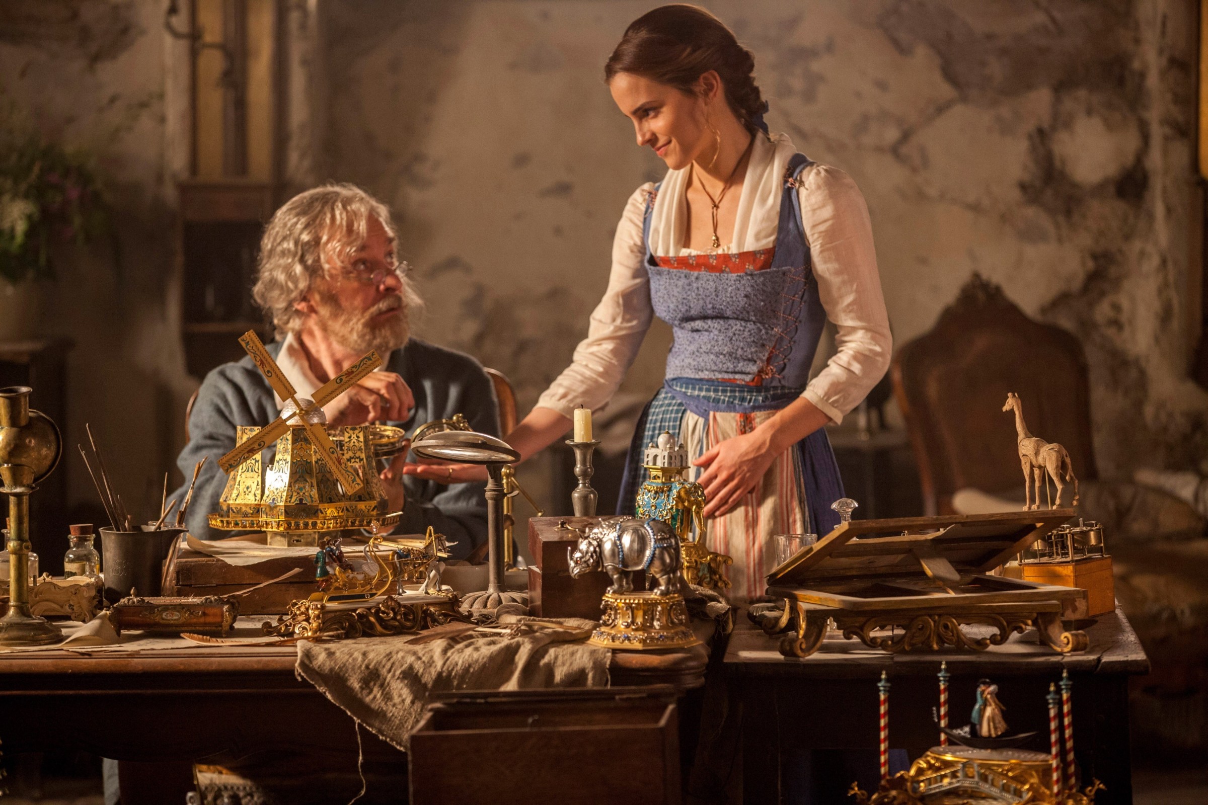 Belle (Emma Watson) smiles down at her father (Kevin Kline) as he sits at his work table, building a little gold toy windmill, in the 2017 live-action Beauty and the Beast.