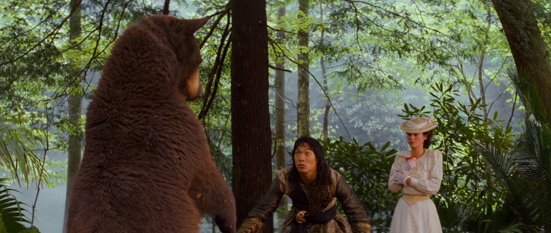 A live bear rears up and towers over a young man in brown and a young woman in a white hat and prim white gown in the 1994 live-action Jungle Book